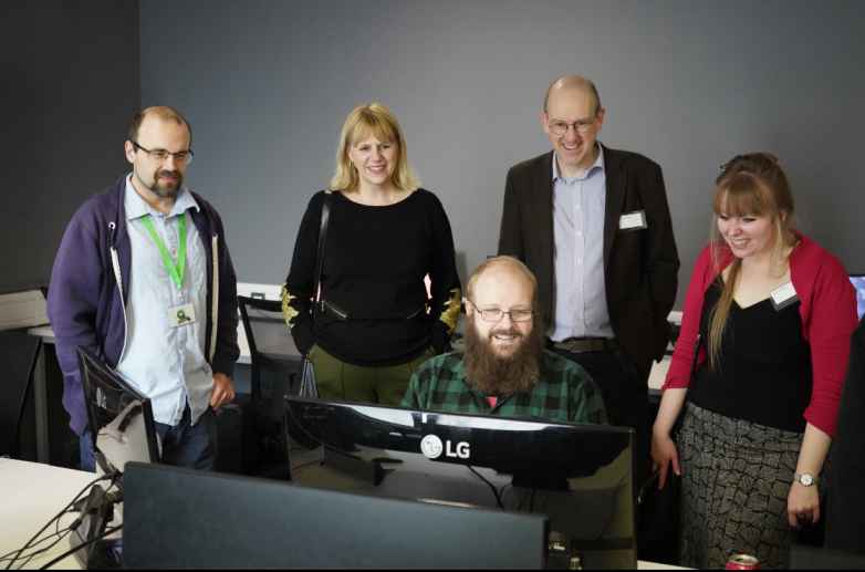 In the foreground a man sits at a computer screen. Standing behind him are four other people who are also looking at the screen. All five people are smiling.